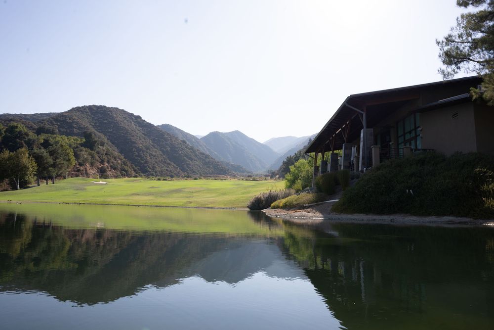 Scenic mountain lake reflects green meadow and rustic building against forested hillside in tranquil valley setting.
