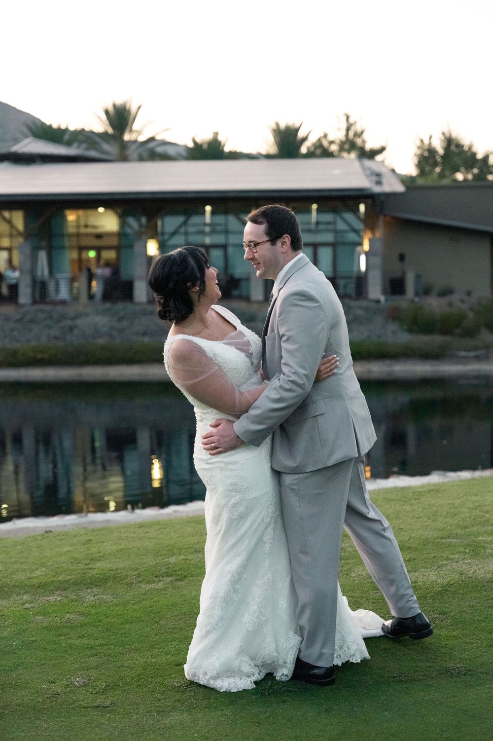 A couple shares their first dance together on a grassy lawn near a lake at sunset, with a modern building in the background.