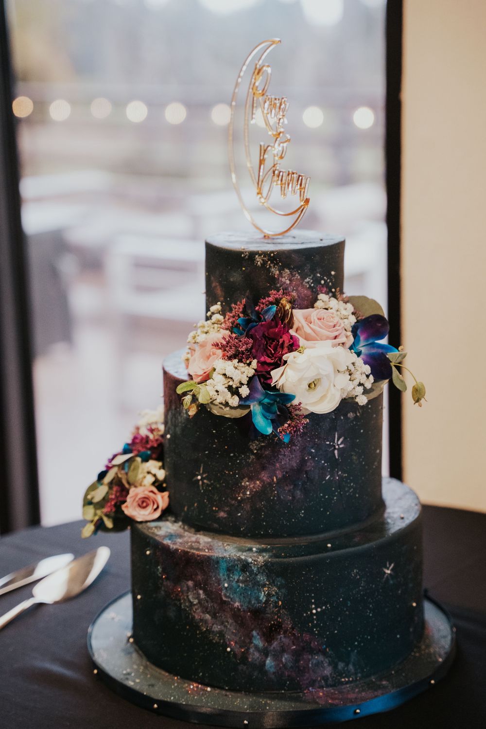 Three-tier black galaxy themed wedding cake decorated with fresh flowers and gold cake topper on dark table.