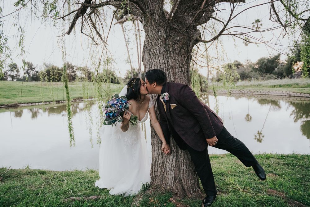 A romantic couple in wedding attire share a tender moment leaning against a willow tree beside a serene pond.