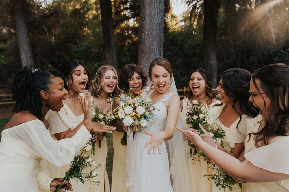 Wedding party in cream colored dresses laughing together while holding white and yellow flower bouquets in outdoor setting.