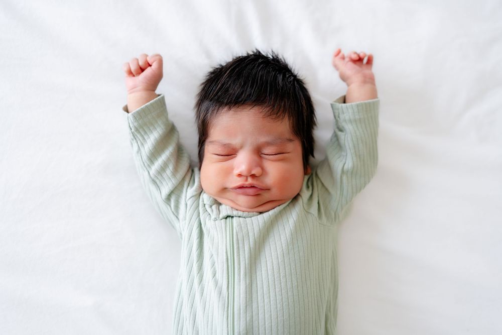 Sleeping newborn baby in mint green ribbed outfit stretches arms up while lying on white bedding.
