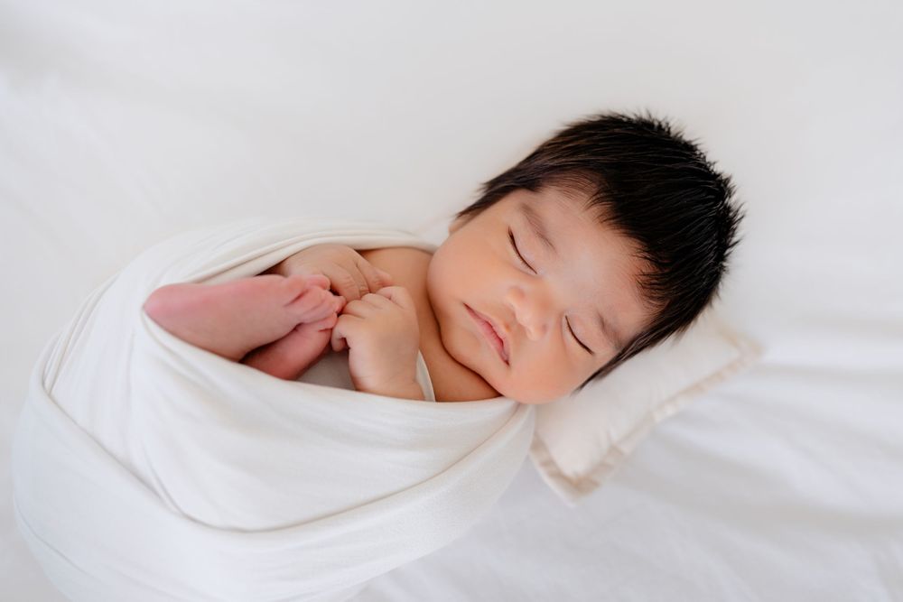 Sweet newborn baby with dark hair sleeping peacefully while swaddled in white.