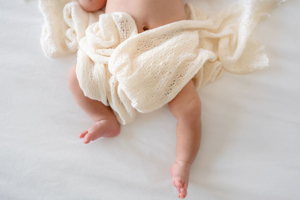 Baby wearing cream-colored lace dress lying on white fabric with tiny feet showing from underneath the dress material.