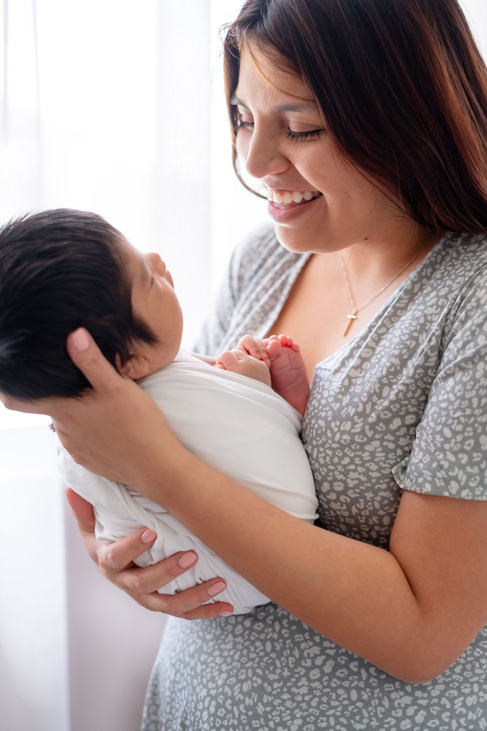 A person in a gray floral dress holds and gazes lovingly at a newborn baby near a window with white curtains.