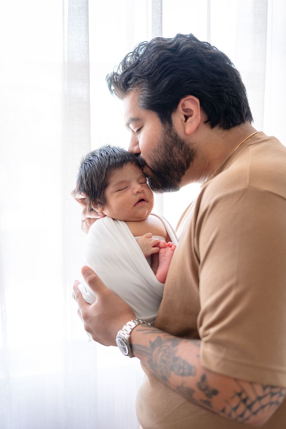 Parent in brown sweater tenderly holding sleeping newborn baby against window light.
