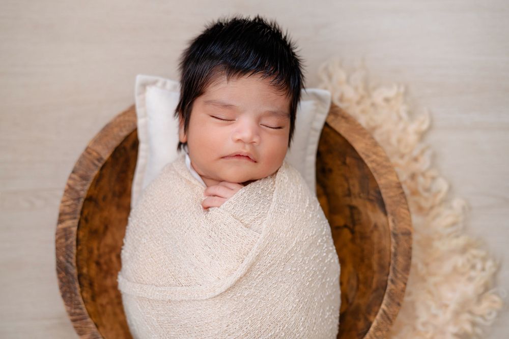 Sleeping newborn wrapped in cream swaddle lying in wooden bowl with textured backdrop.
