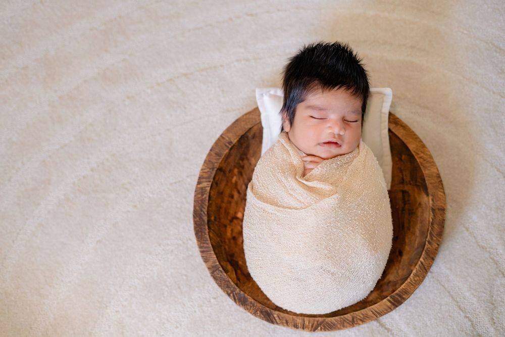Swaddled sleeping newborn resting in round wooden bowl on white background.