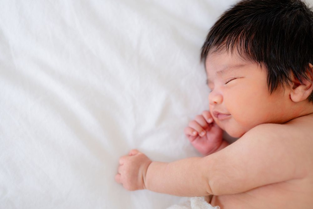 Peaceful sleeping baby lying on white bedding with dark hair and tiny outstretched arm.
