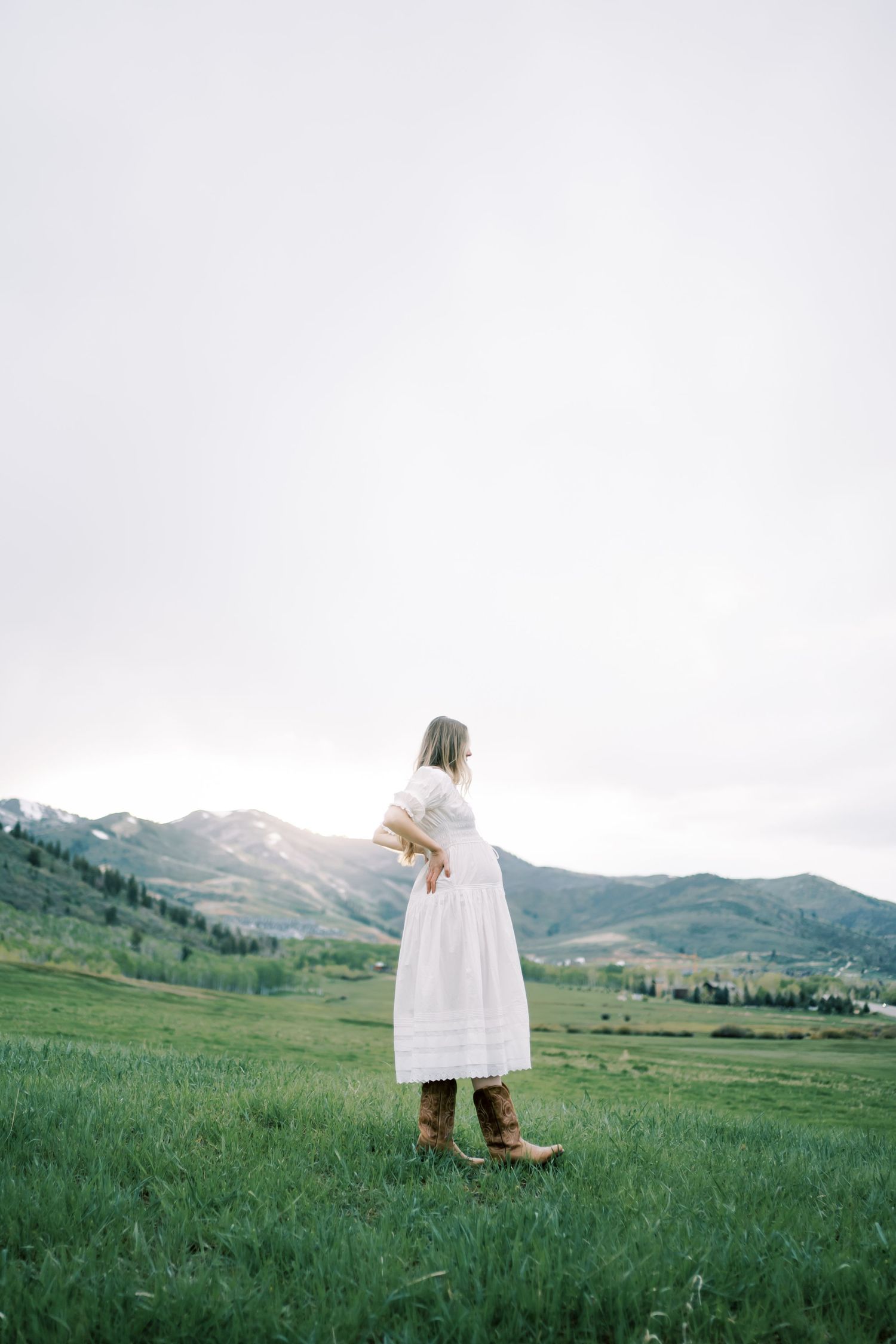 Person in a white dress stands in a grassy meadow with mountain ranges stretching across the horizon.