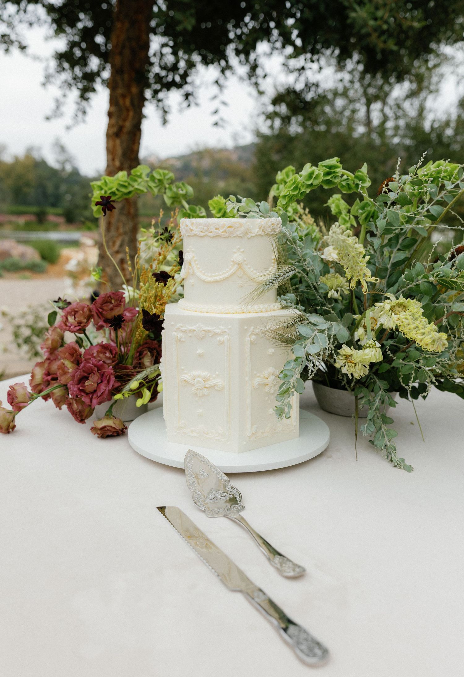 An elegant white two-tier wedding cake decorated with delicate scallops and surrounded by fresh floral arrangements.