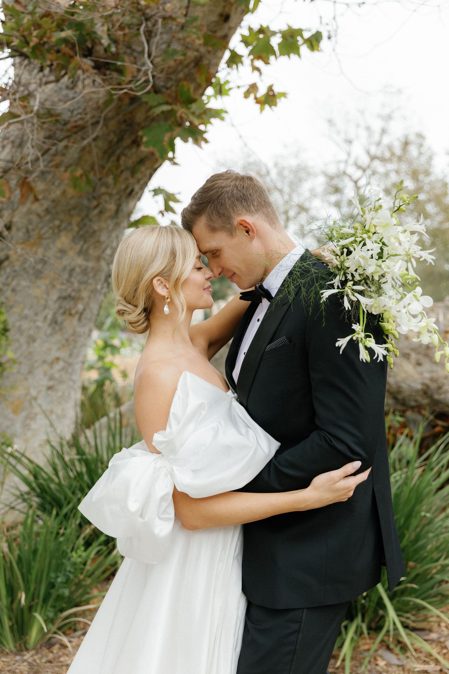 A romantic moment between newlyweds embracing outdoors with a large tree and foliage in the background.