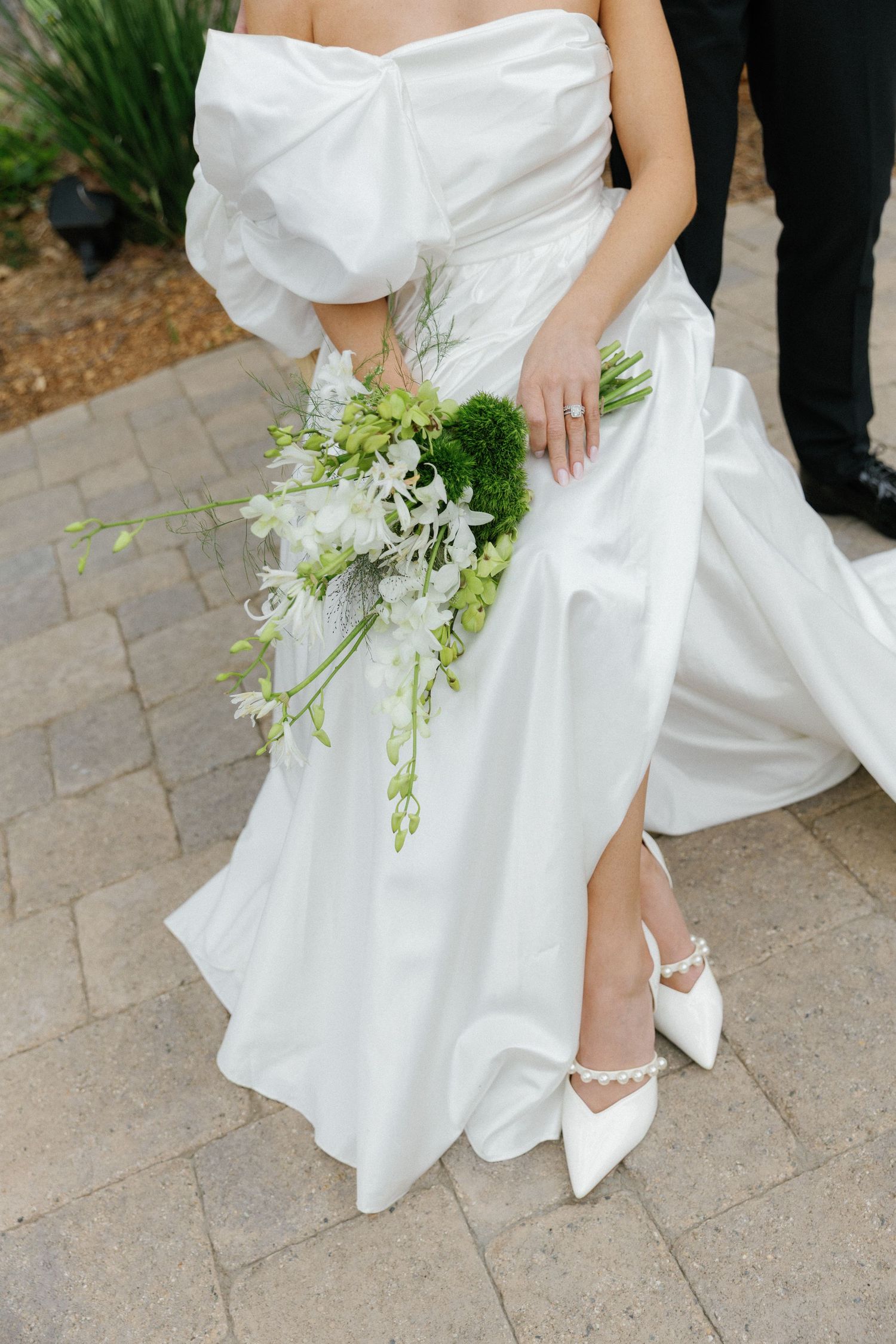 Close-up view of a white wedding dress with a flowing skirt, pointed bridal shoes and a green and white bouquet.
