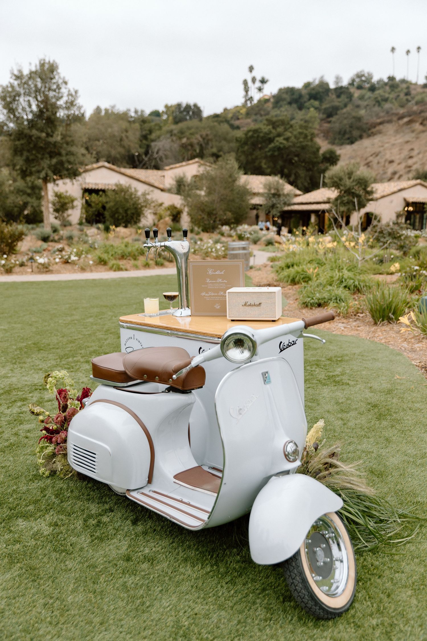 A vintage white Vespa scooter with brown leather seat parked on grass with buildings and hills in the background.