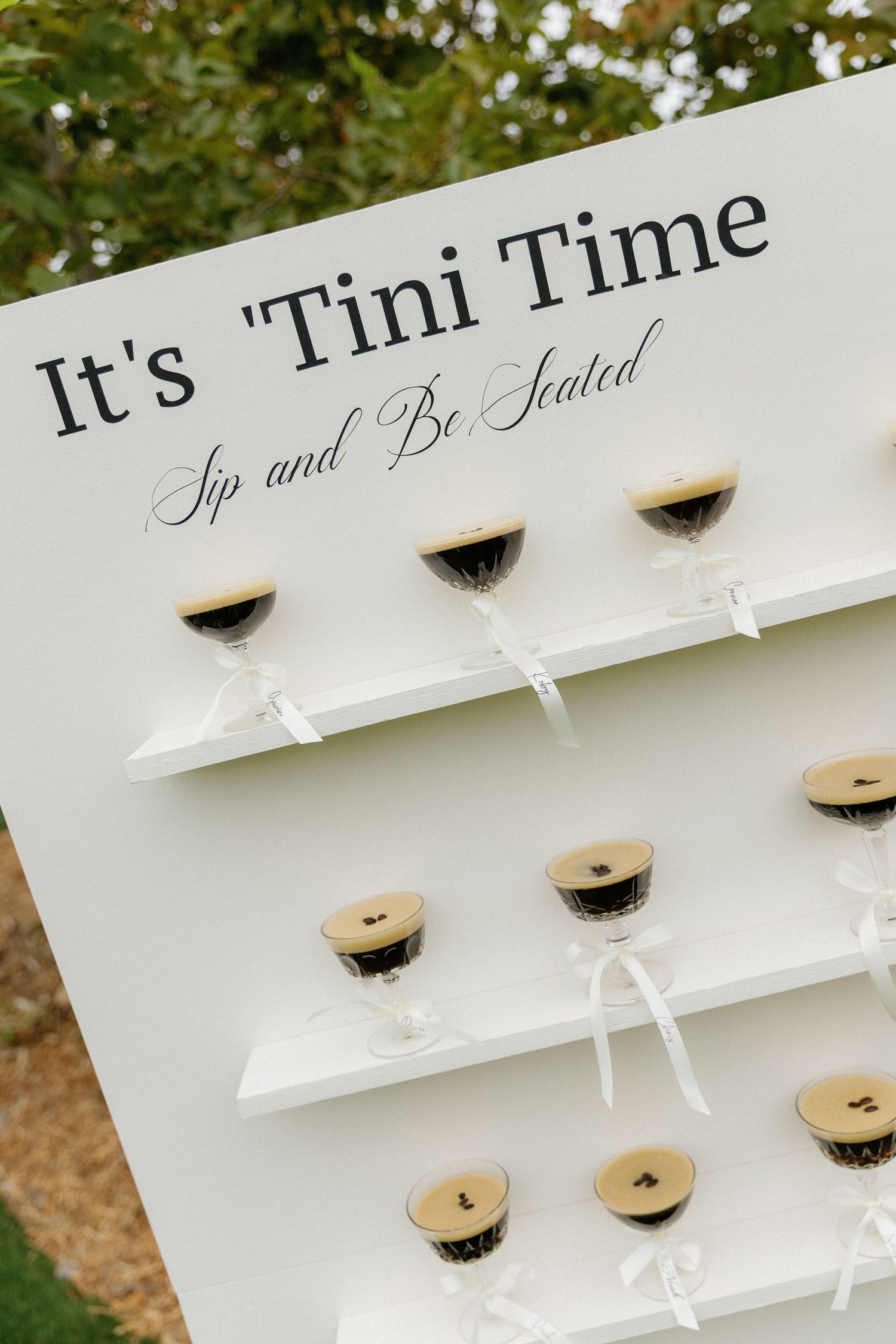 A white display wall with 'It's Tini Time' text and shelves holding mini martini glass party favors.