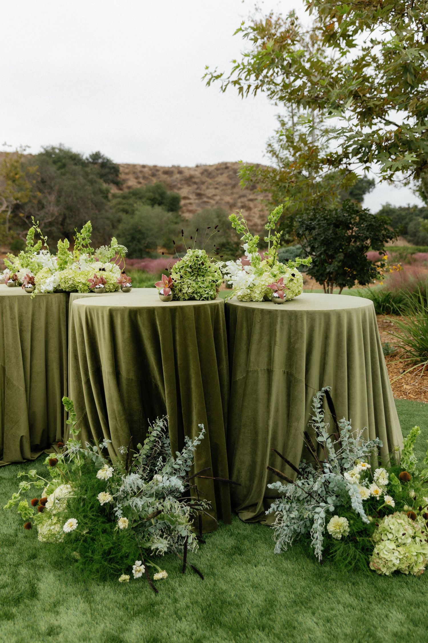 Elegant wedding cocktail tables with olive green velvet tablecloths adorned with floral arrangements in an outdoor garden setting.
