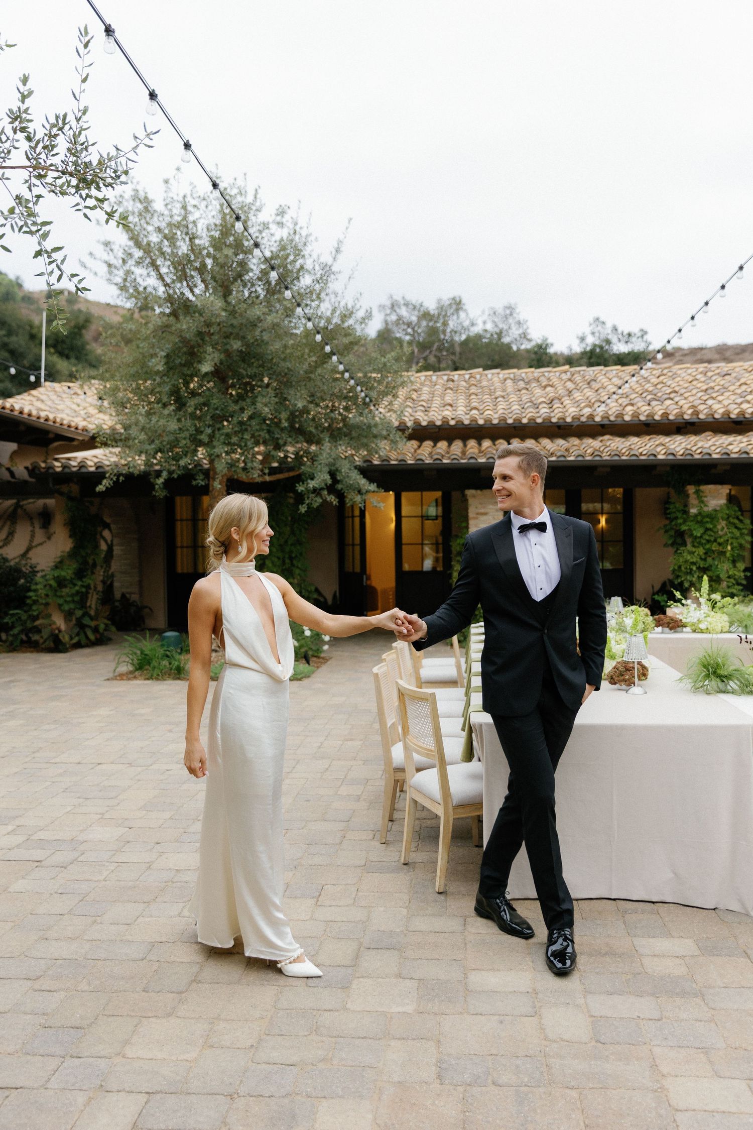 Elegant couple in formal attire sharing a moment in a Spanish-style courtyard with terracotta roof and string lights.