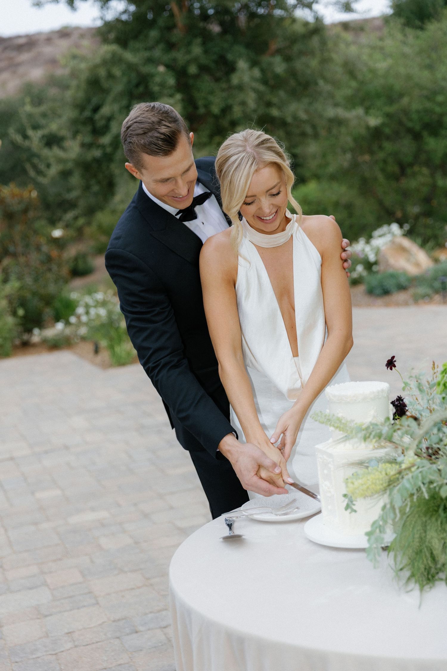 A couple in formal attire together cutting their white wedding cake at an outdoor garden reception.