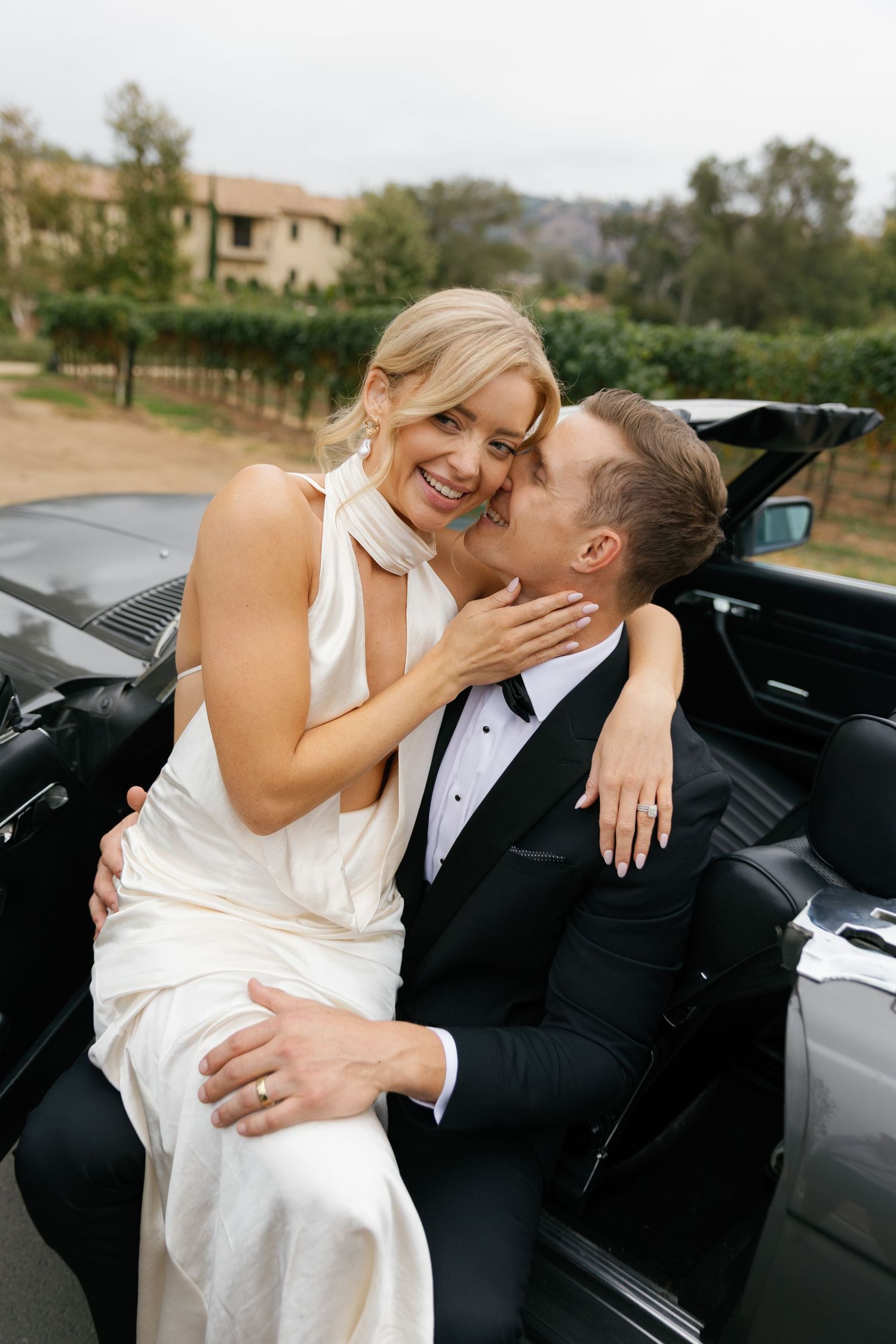 A romantic moment between newlyweds sharing laughter while sitting in a classic car after their wedding ceremony.