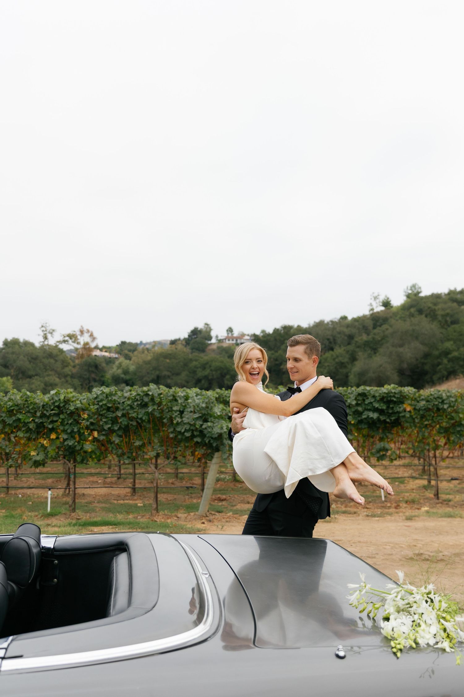 Wedding couple embraces near a convertible car parked beside a vineyard in a romantic outdoor setting.