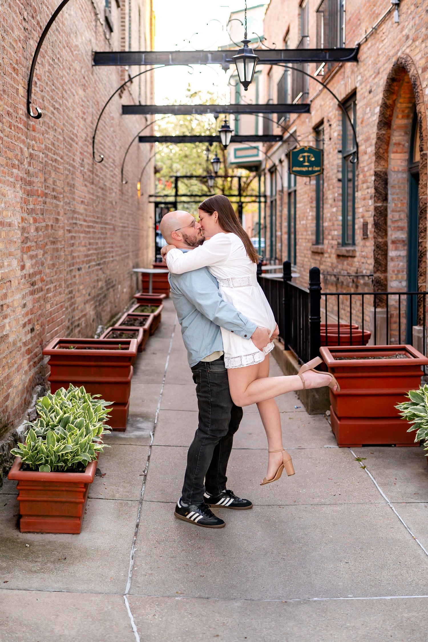 A romantic moment between a couple embracing in an arched brick alleyway with potted plants and decorative lighting.
