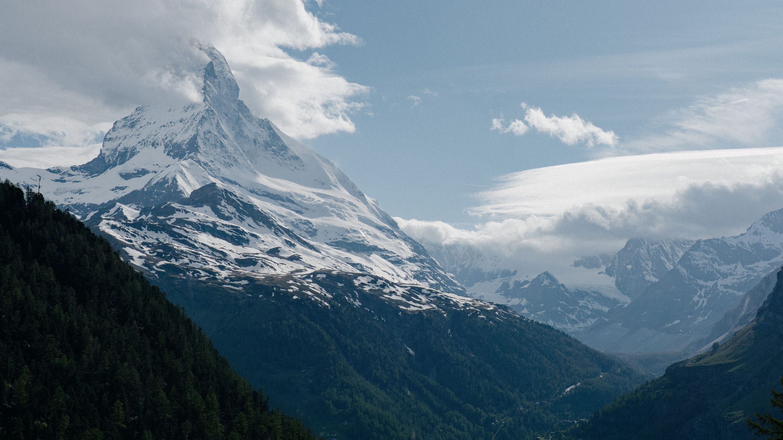 Snow-capped Matterhorn peak towers over a scenic Alpine valley in Switzerland under partly cloudy skies.