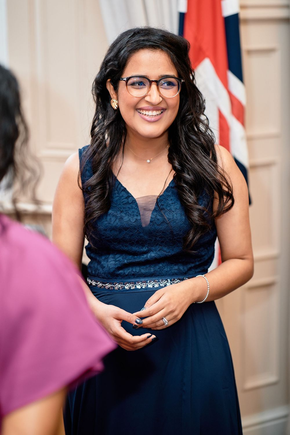 A person in glasses and a navy blue formal dress stands smiling while conversing at an event with flags in the background.