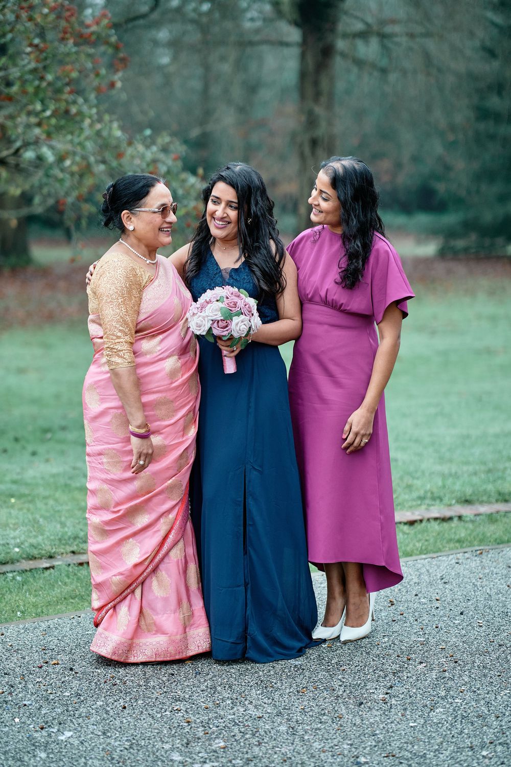 Three women in elegant dresses gather outdoors, wearing pink, navy and purple, with one holding a floral bouquet.