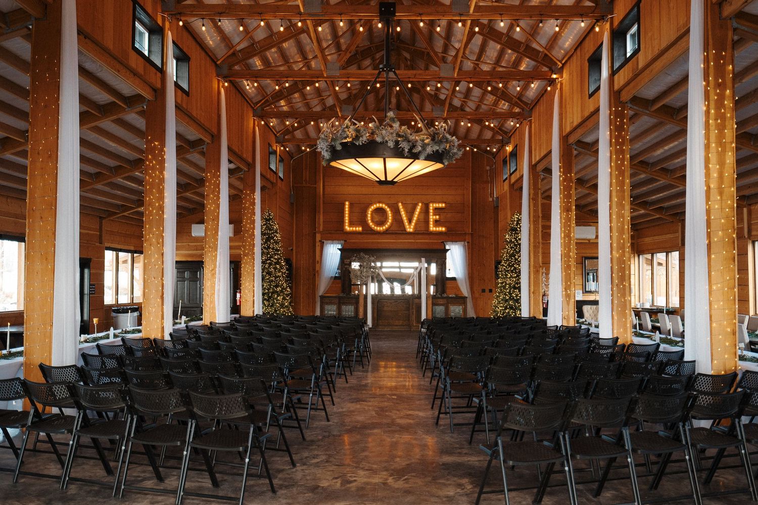 Rustic indoor wedding venue with wooden beams, string lights, rows of black chairs, and illuminated LOVE sign at the altar.