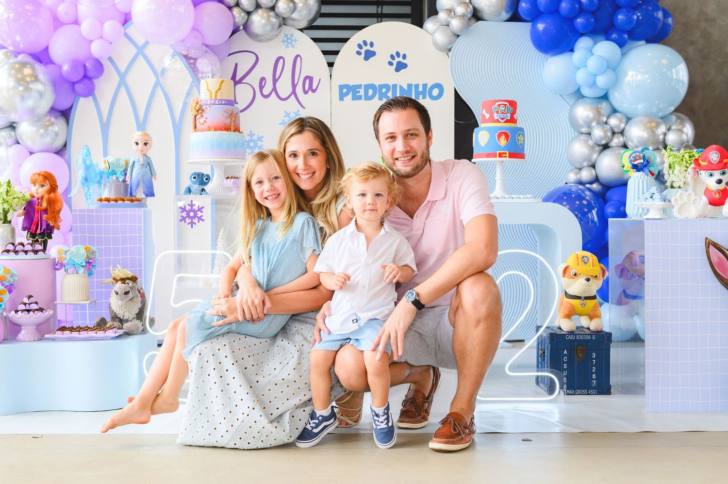 A family poses in front of a festive blue and purple balloon arch and decorative backdrop at a birthday celebration.