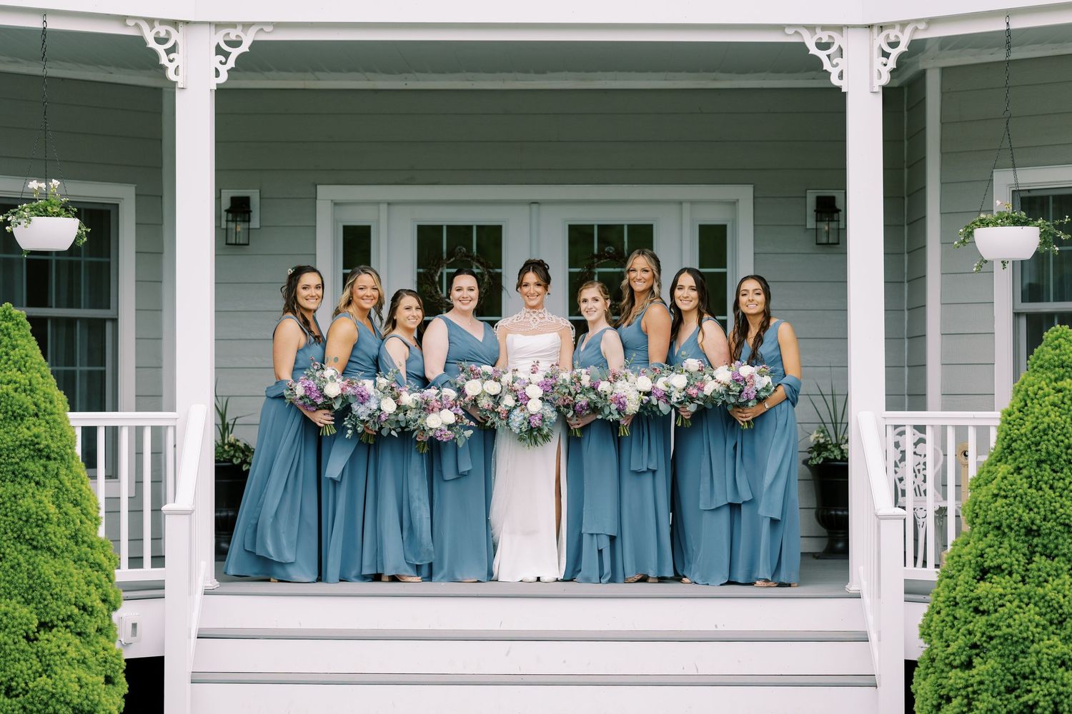 Wedding party in blue bridesmaid dresses and white wedding gown pose on front porch of classic white house at Windham Manor.