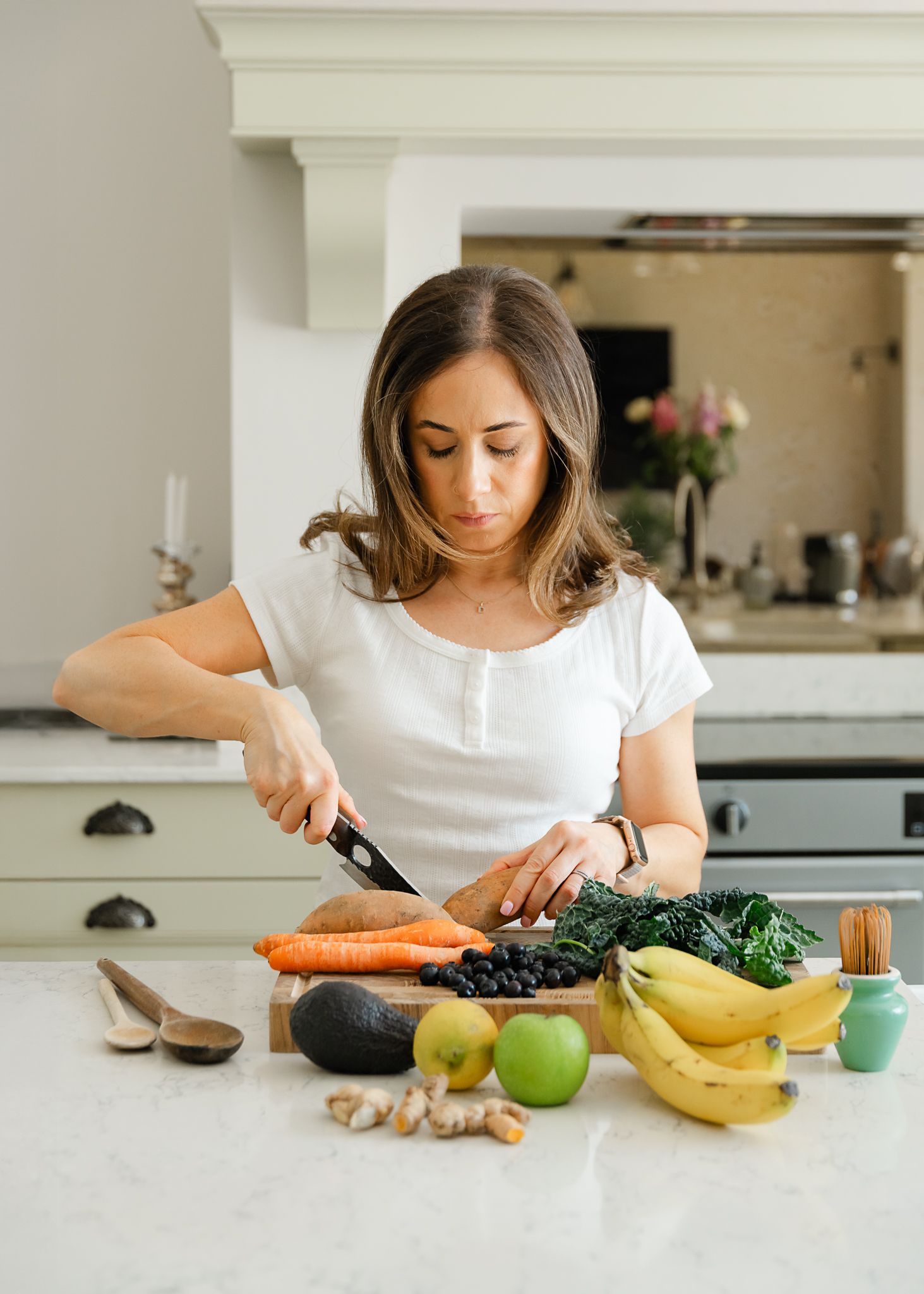 Person chopping fresh vegetables and preparing healthy whole foods on white kitchen counter with bananas and apples.