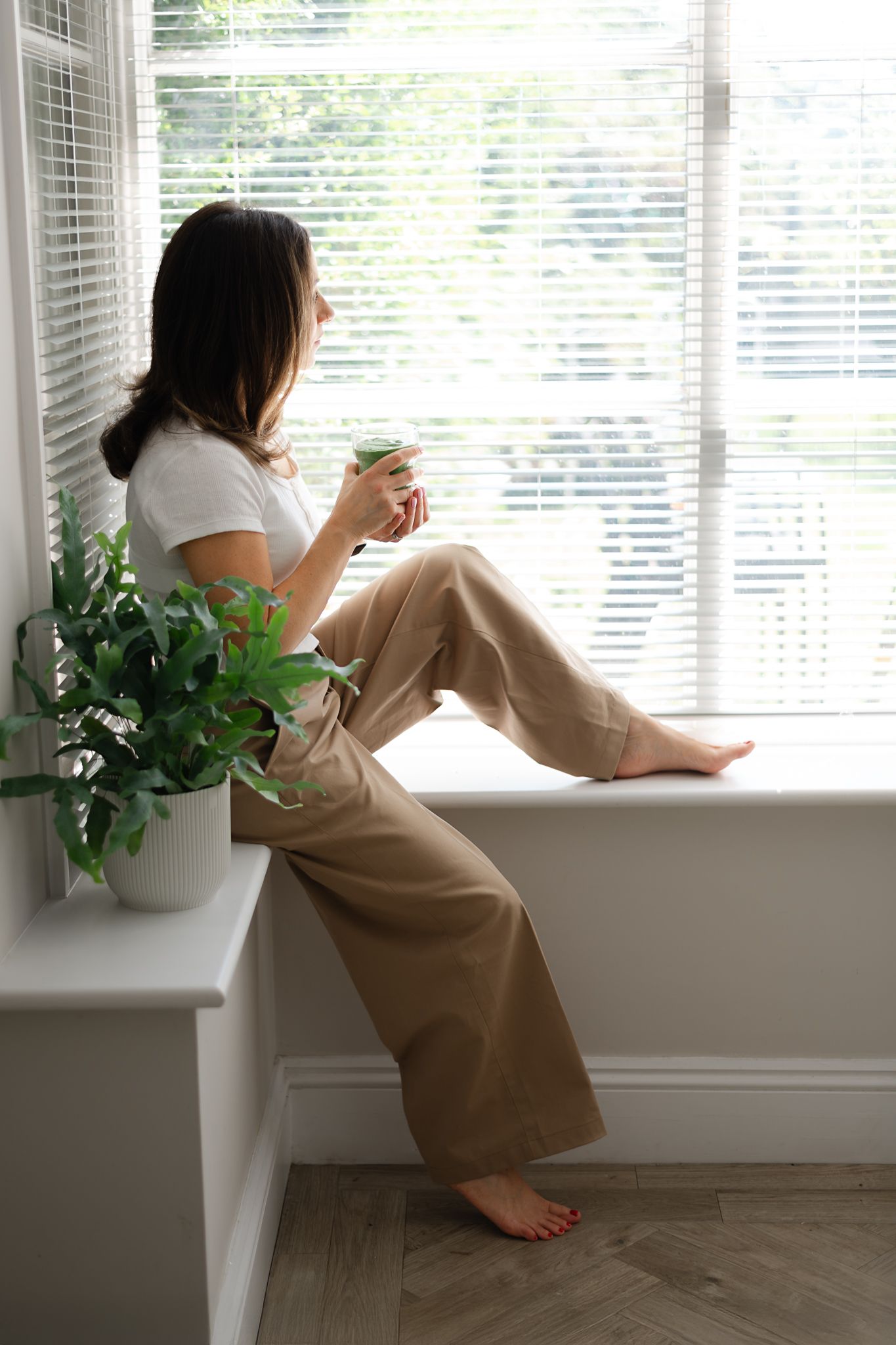 Relaxing by window with coffee in white shirt and beige pants next to potted plant on sunny morning.