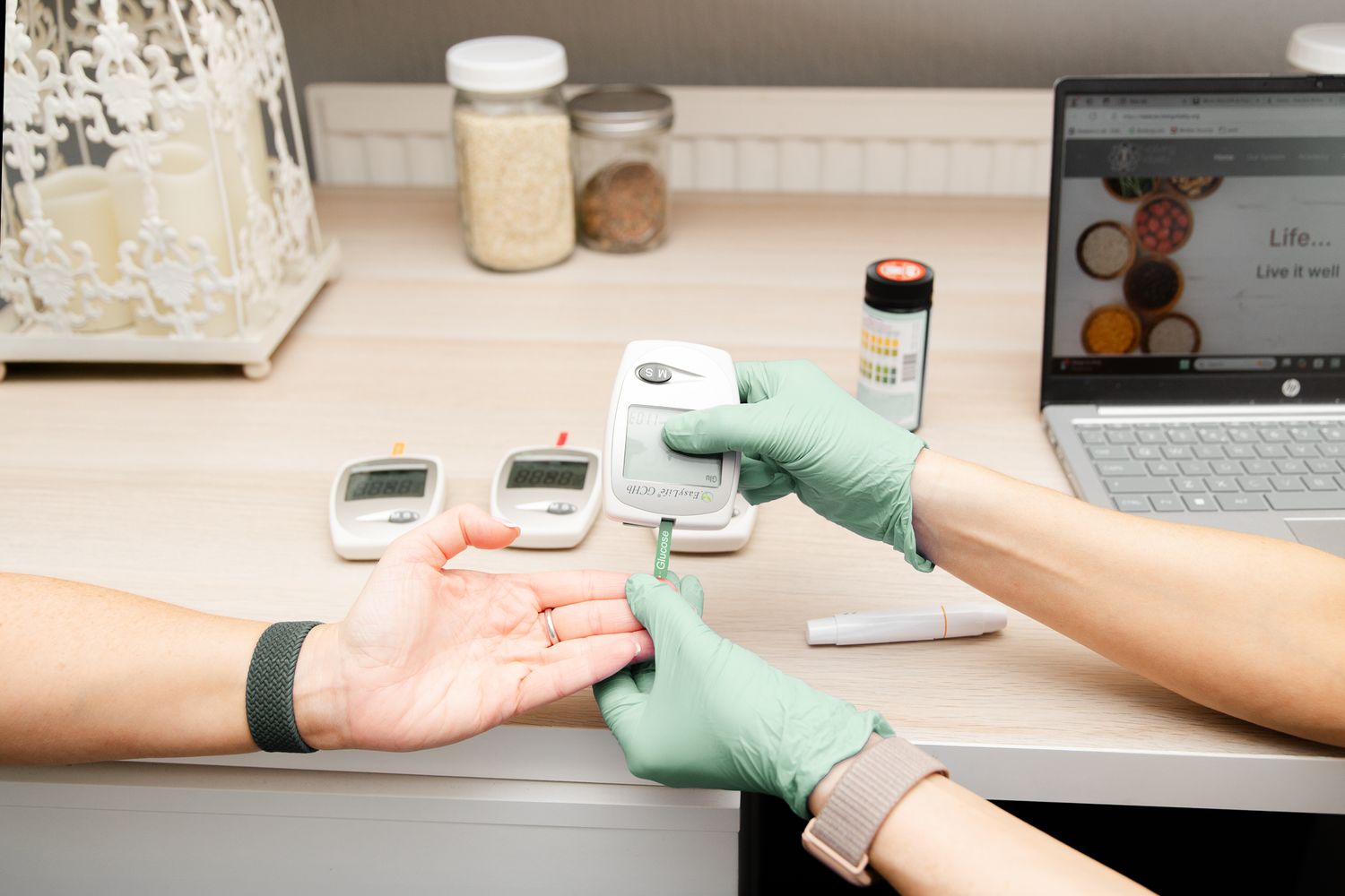 Healthcare provider wearing mint green gloves performs glucose test on patient's finger while laptop displays data.