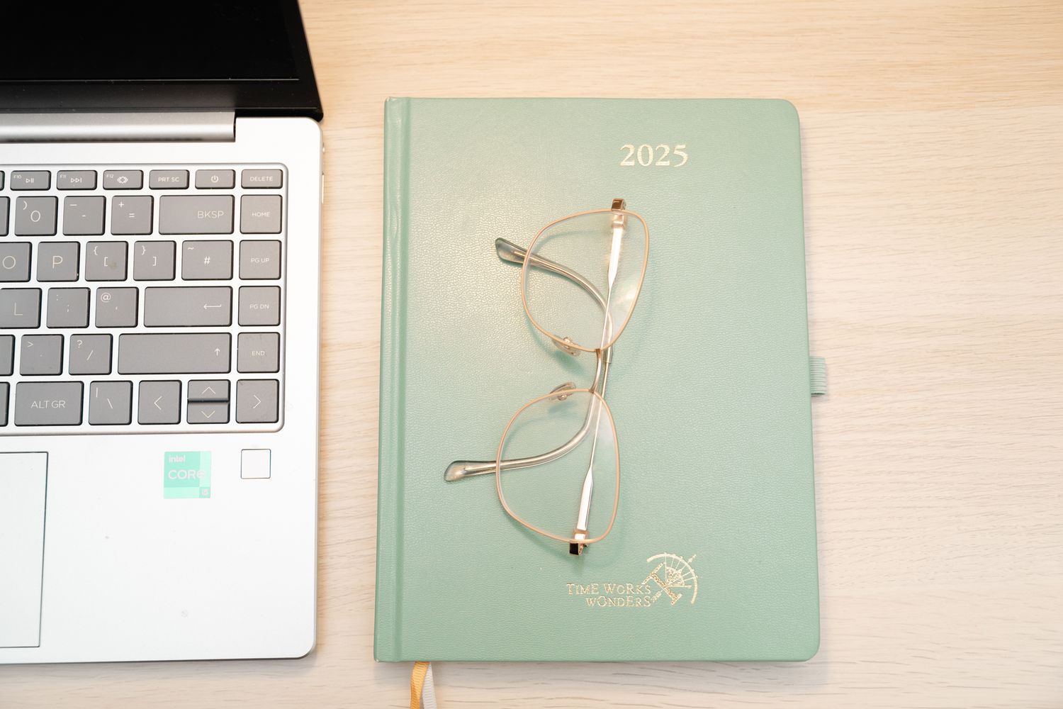 A laptop keyboard next to a mint green 2023 planner with gold-rimmed glasses resting on top on a wooden desk surface.