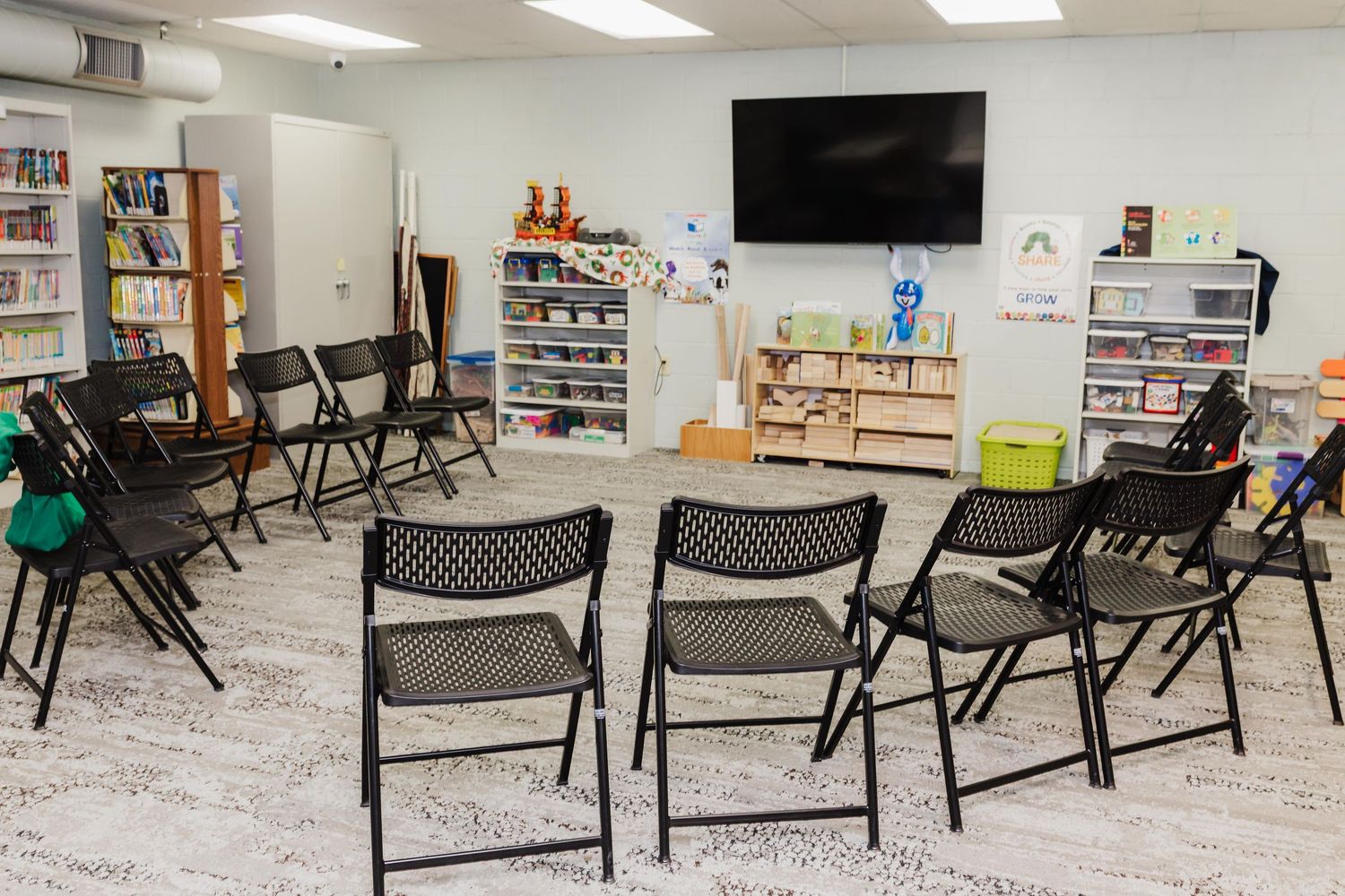 Empty folding chairs arranged in a circle face a bookshelf and TV in a local library storytime space.