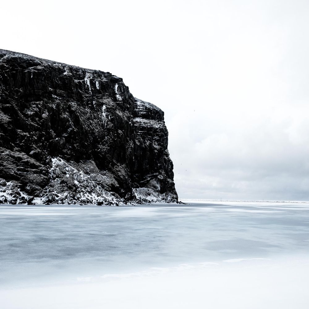 A dramatic black and white coastal landscape featuring a snow-dusted cliff face rising above misty waters.