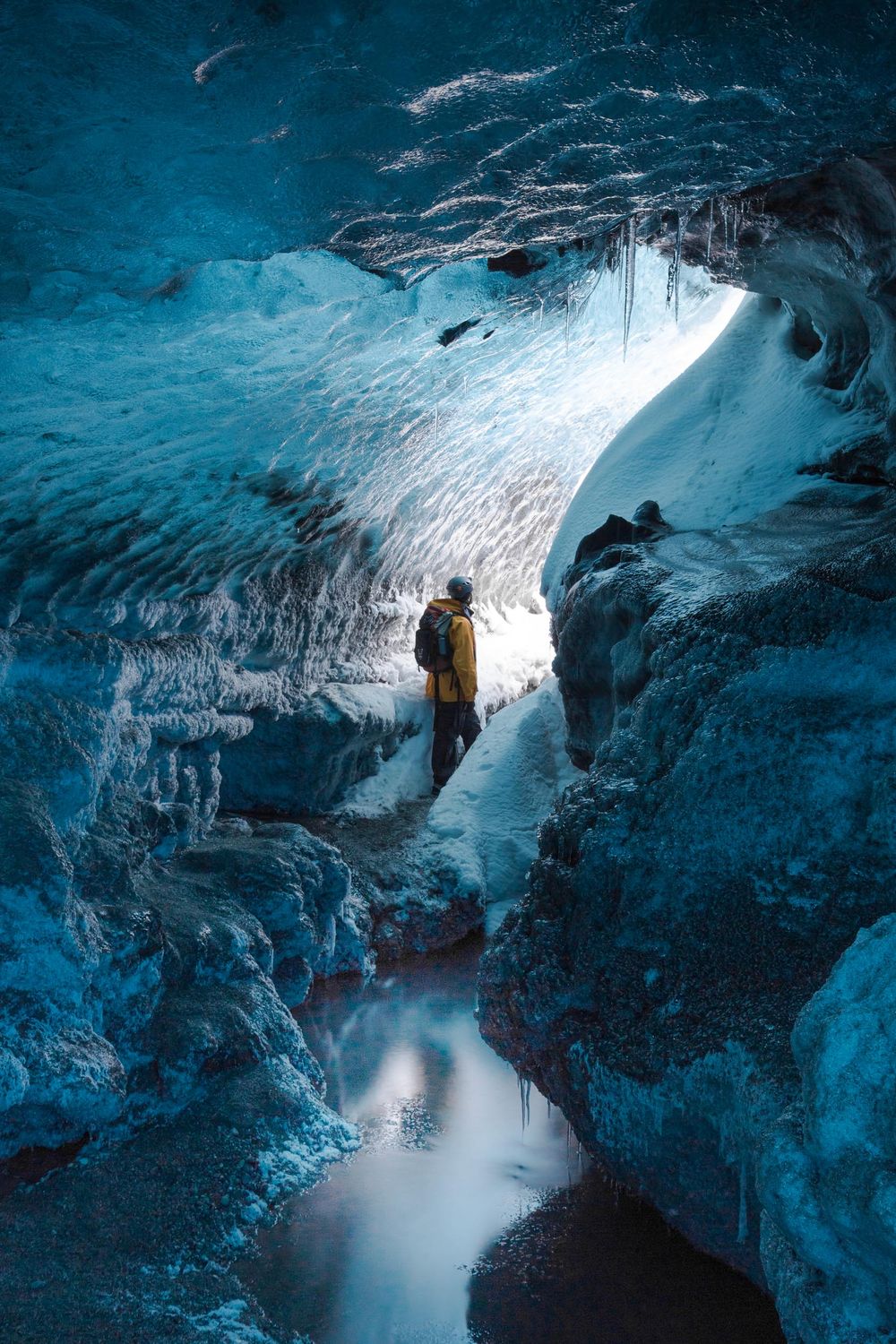 Explorer stands inside a stunning blue ice cave in Iceland with ethereal light streaming through glacial formations.