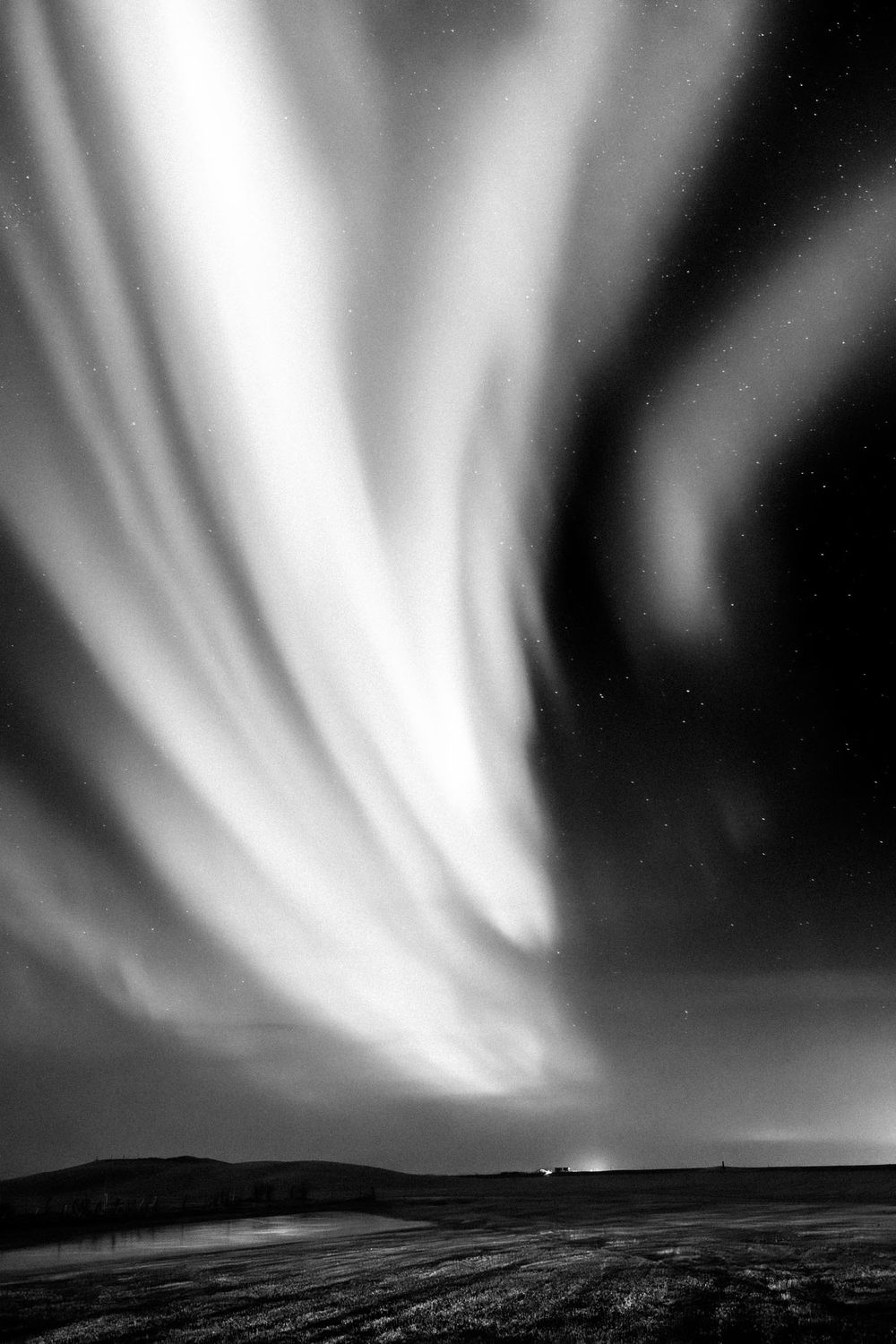 Dramatic black and white photo of the Northern Lights swirling across a dark night sky over a barren landscape.