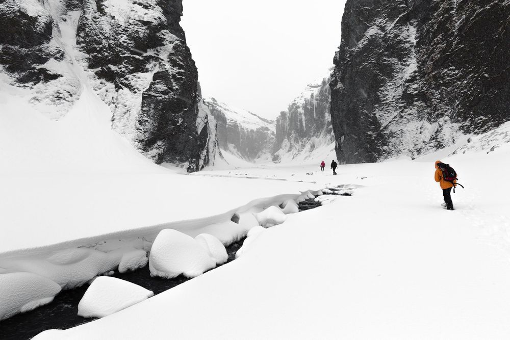 Winter hikers trek through a snowy canyon with icy rocks and cliffs towering on both sides.