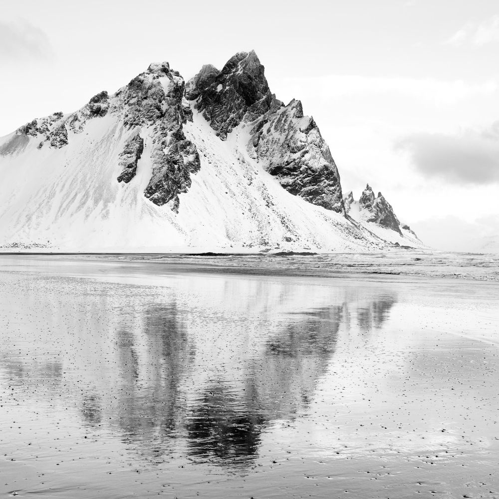 A dramatic black and white mountain landscape reflected in calm waters at Stokksnes beach in Iceland.