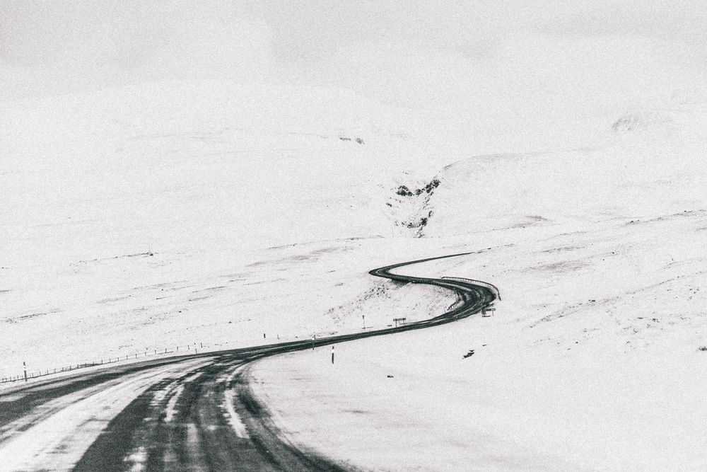 A curving winding road cuts through a stark snow-covered landscape under an overcast winter sky.
