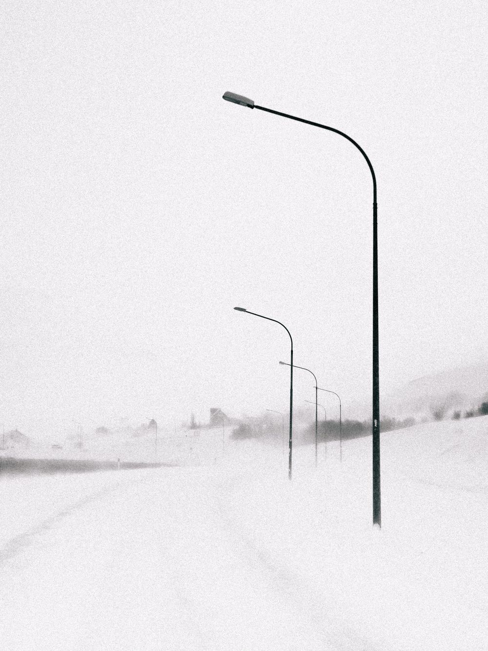Minimalist black and white photograph of street lamps along a snowy road creating stark contrast against light sky.