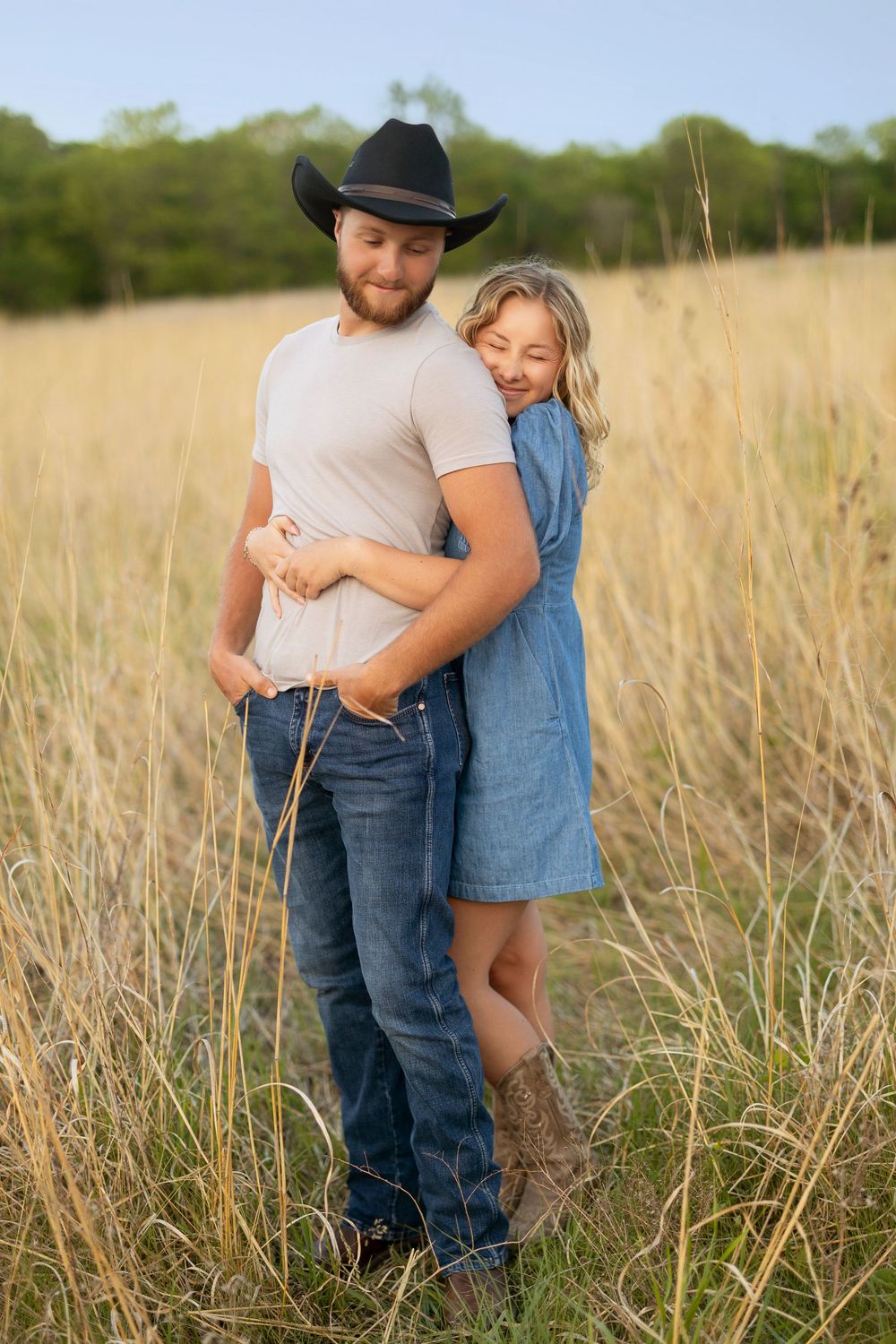 A couple embrace in a sun-lit field of tall grass during golden hour, with the person in denim leaning against their partner.