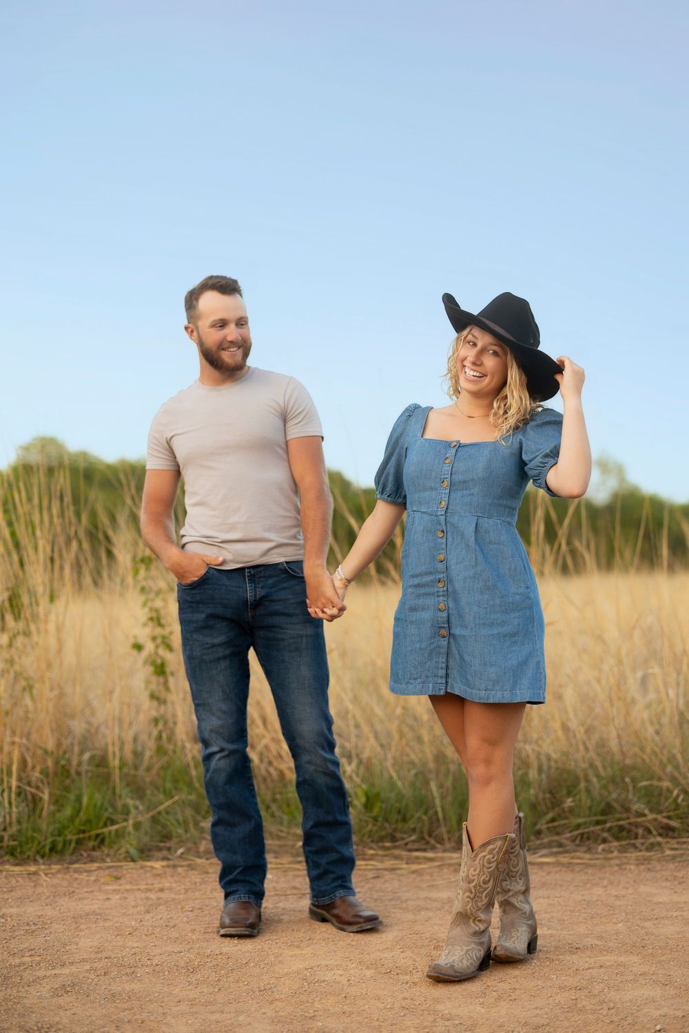 A couple in casual attire holds hands while standing in a golden field during sunset or sunrise.