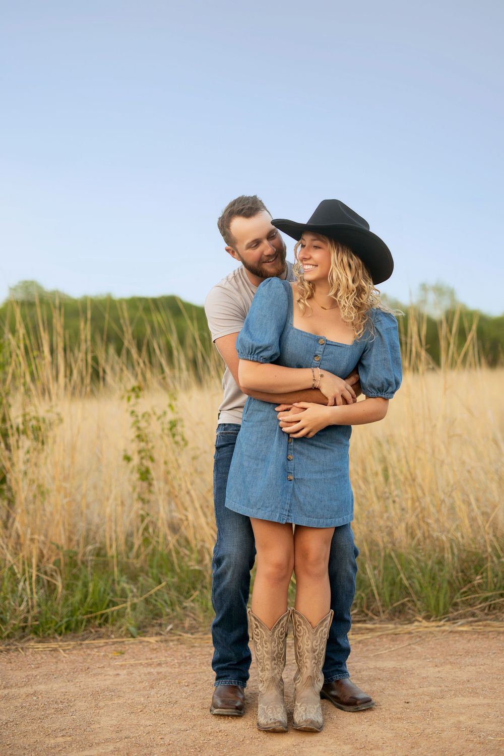 A couple embraces on a dirt path surrounded by tall grass during golden hour, wearing cowboy boots and western attire.