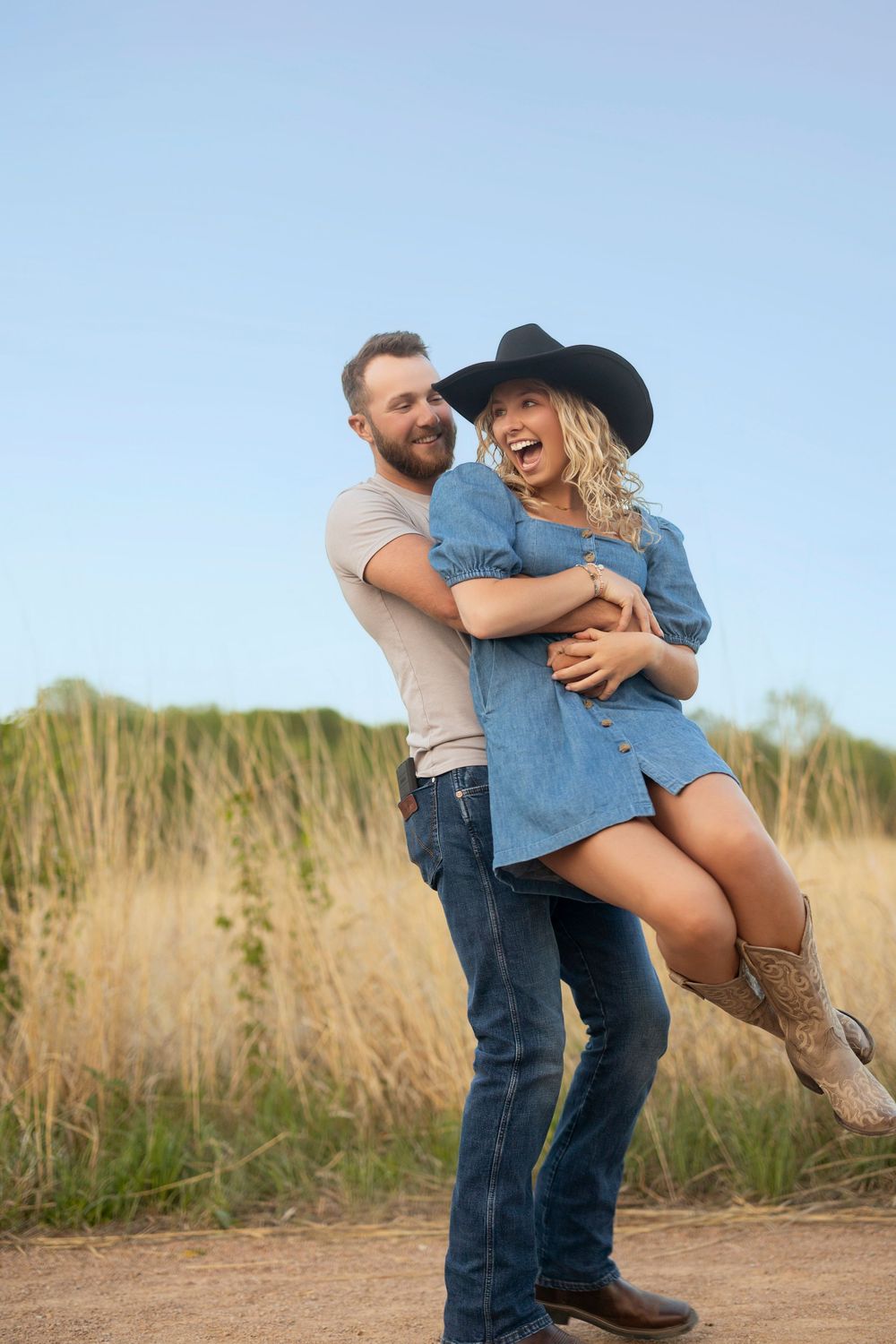 A couple embraces playfully in a grassy field at sunset, with her wearing a cowboy hat and denim shirt against a blue sky.