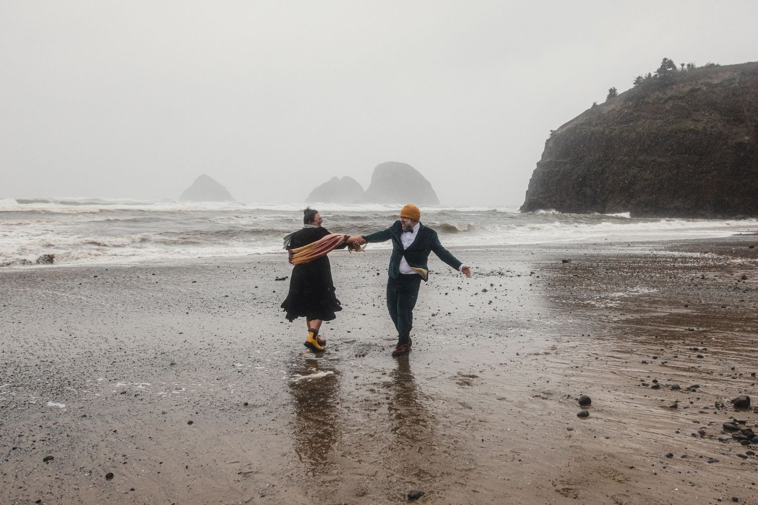 Two people in wedding attire stand on a misty rocky beach shore during a moody coastal photoshoot.