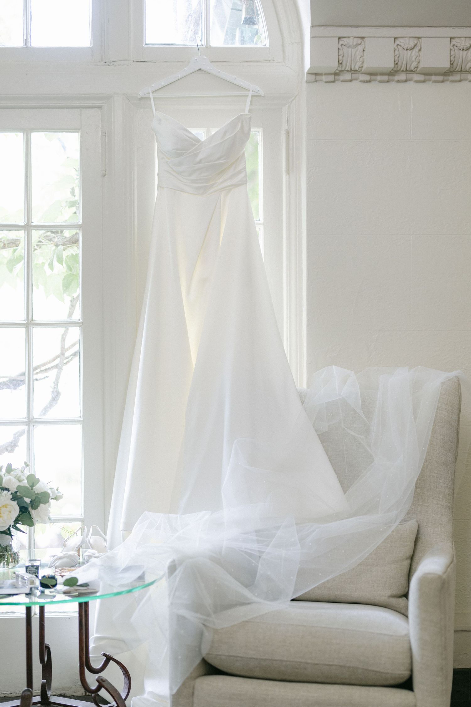 An elegant white wedding dress hangs by a bright window next to a cream-colored armchair and glass side table.