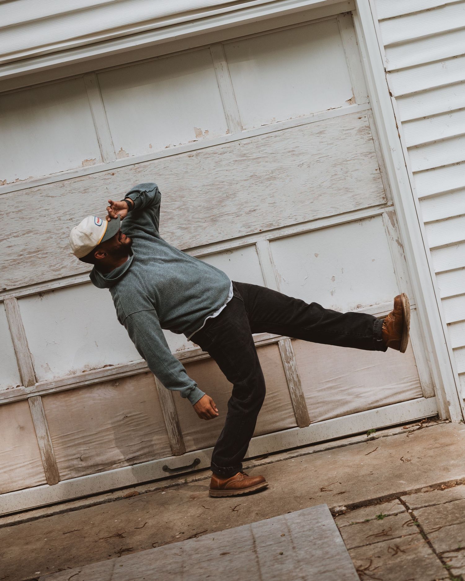 Person in dark clothing and boots balancing on one leg against a white wall, creating a dynamic pose for photography.